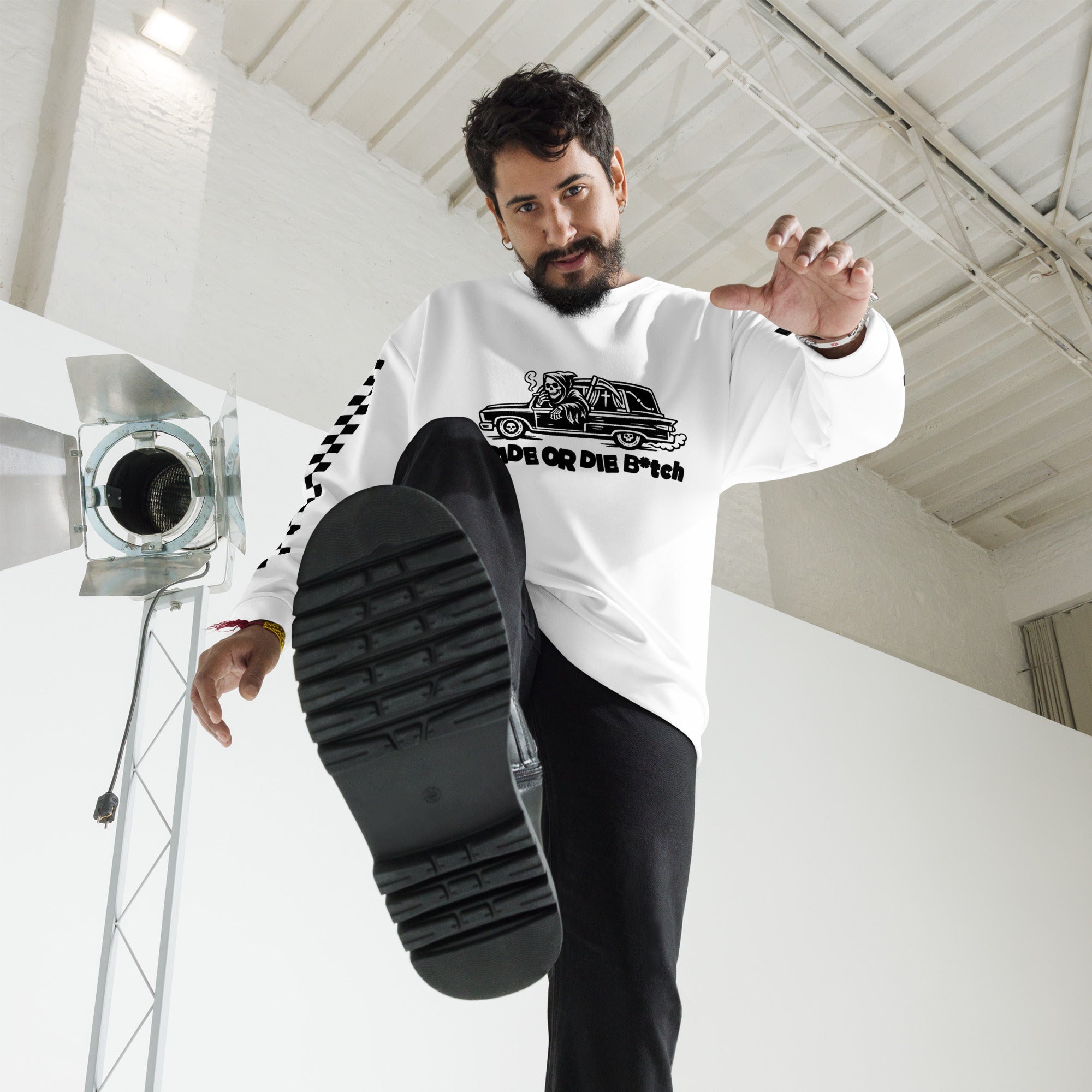 Man in a white sweatshirt with a graphic design, kicking his black sneaker in a studio setting.